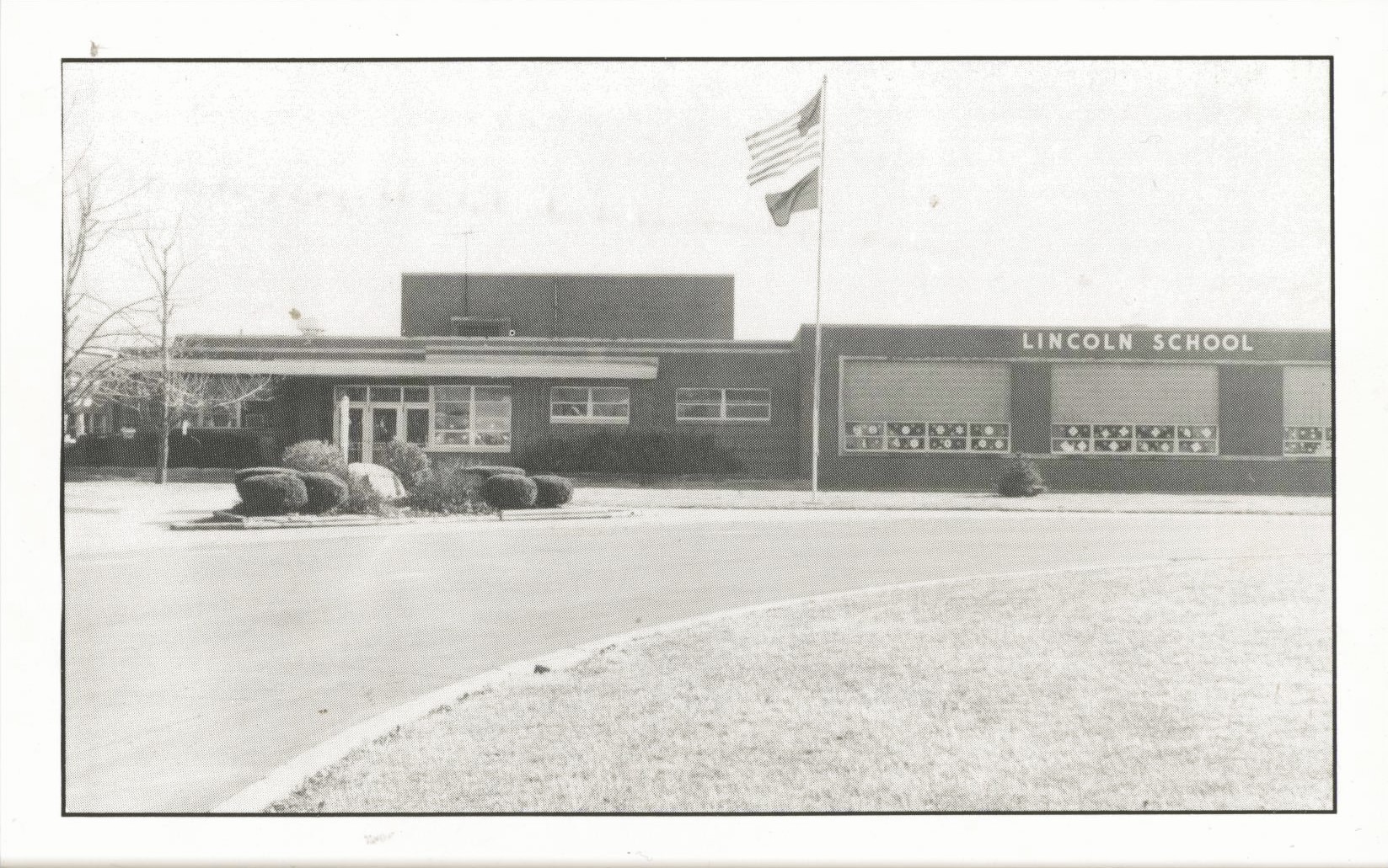 Lincoln Elementary stands behind an American flag flying in the breeze. The circular drive is shown in front of the building.